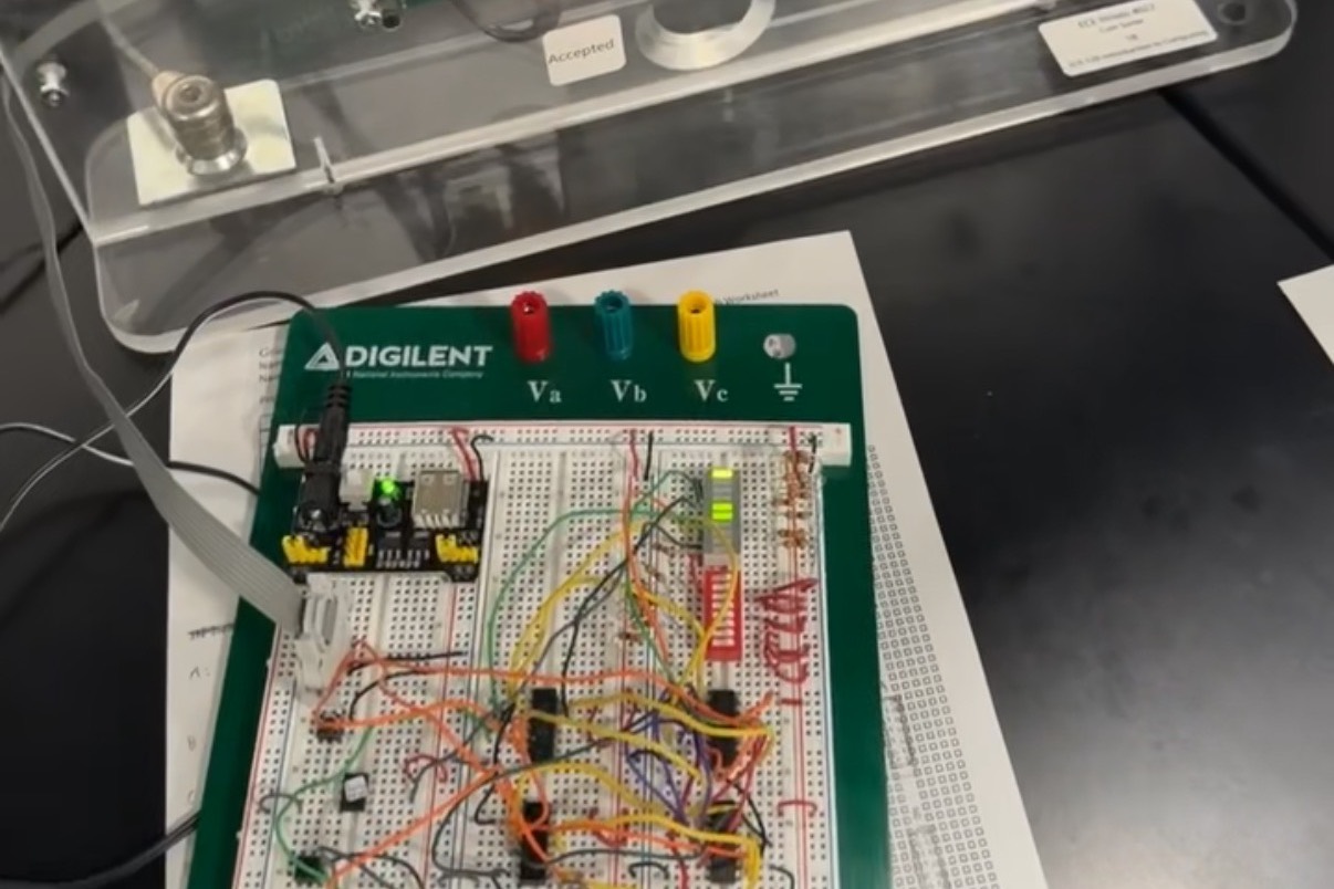 The vending machine controller wired on a breadboard with logic ICs and jumper wires, connected by ribbon cable to the acrylic vending machine prototype behind it.